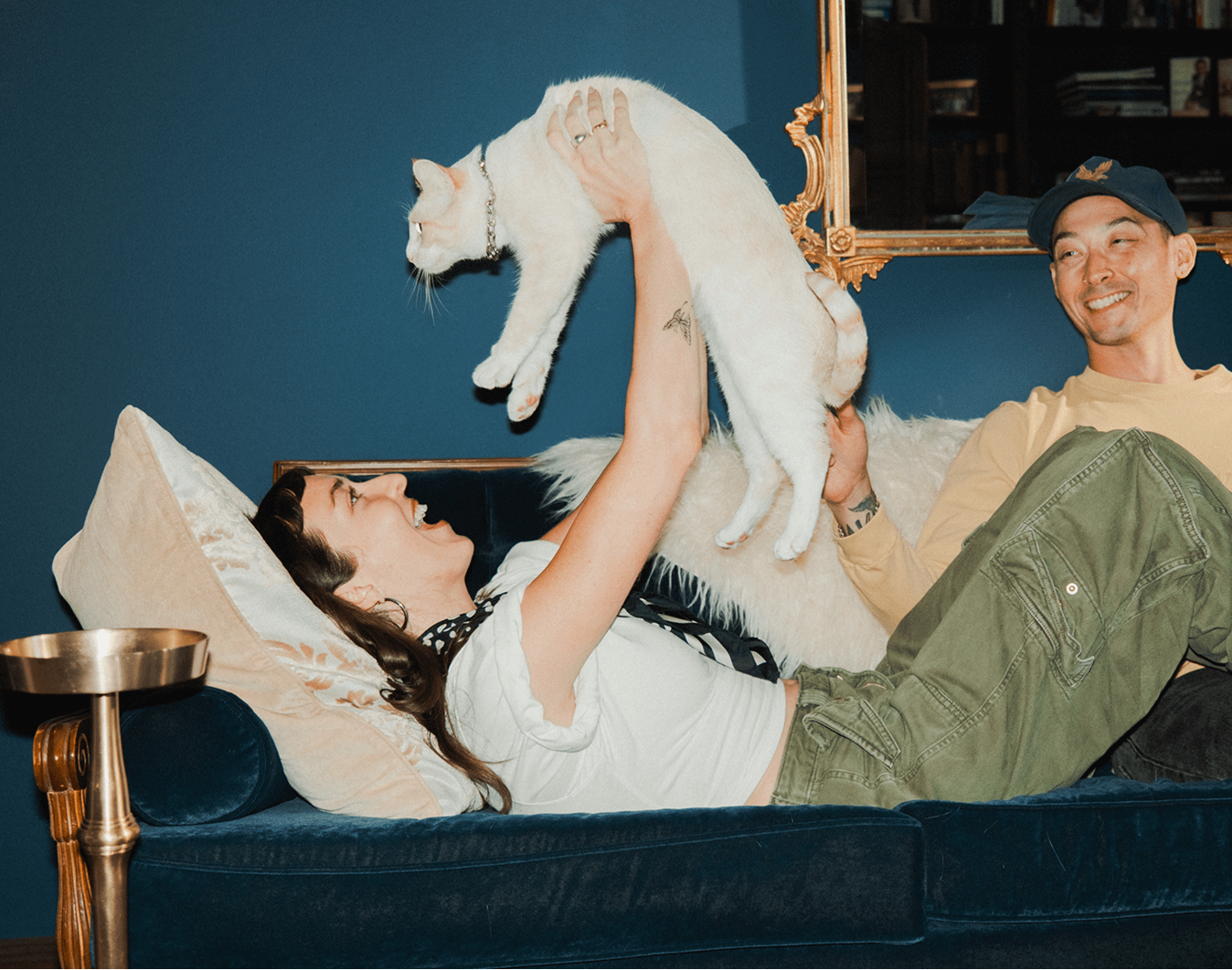 woman laughing lying on couch holding up white cat next to smiling man