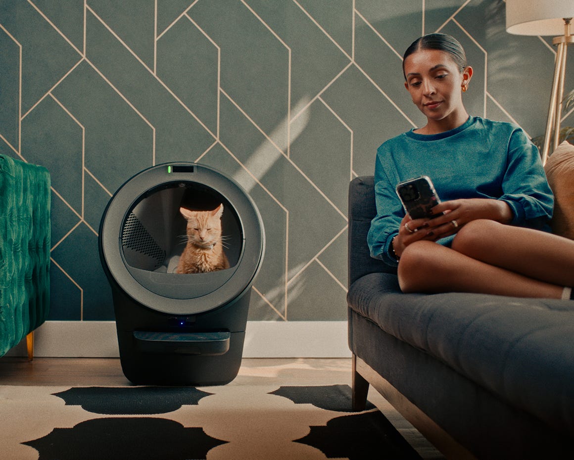 Woman on couch reviewing Whisker app while cat is in Litter-Robot 5