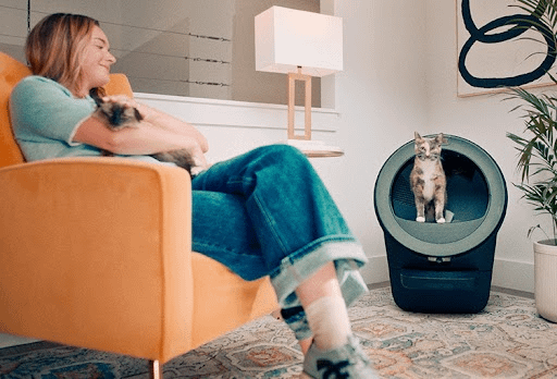 An image of a woman sitting on a chair, petting a cat. There’s a black Litter-Robot 5 next to them with another cat exiting.